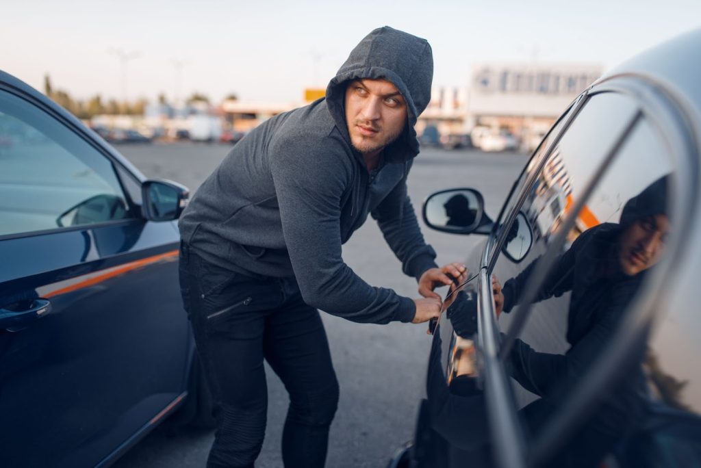 A suspicious individual in a gray hoodie attempting to break into a car in a parking lot, illustrating the risk of auto theft.