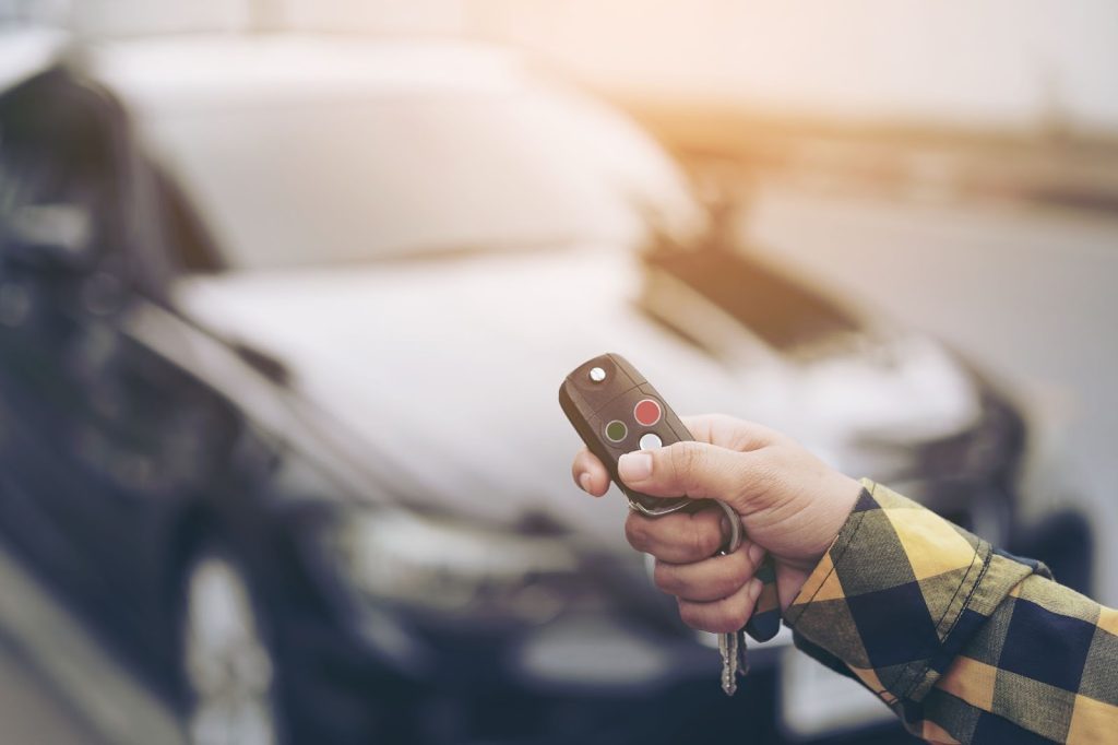 Close-up of a hand holding a car key fob, ready to activate the vehicle's security system, with a blurred car in the background.