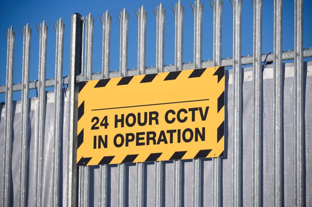 Security fencing with 24-hour CCTV warning sign in a parking lot, crucial for deterring unauthorized access.