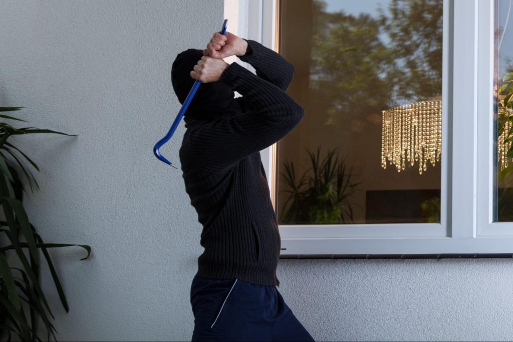 Masked burglar using a crowbar to break into a house through a window.
